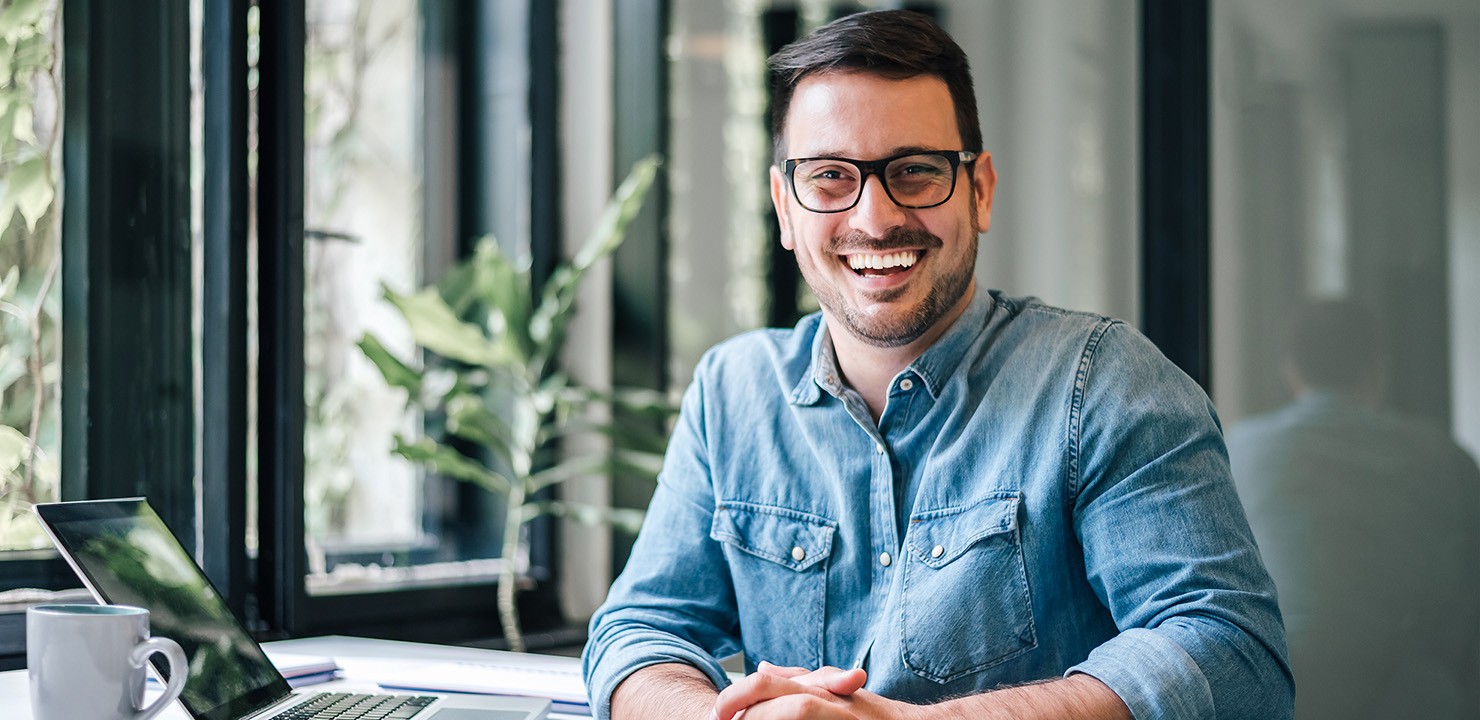 Man in denim shirt sitting at desk with laptop smiling.