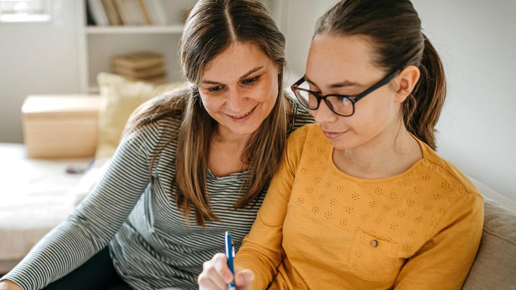 Mother and daughter sitting next to each other on sofa planning college campus tour.