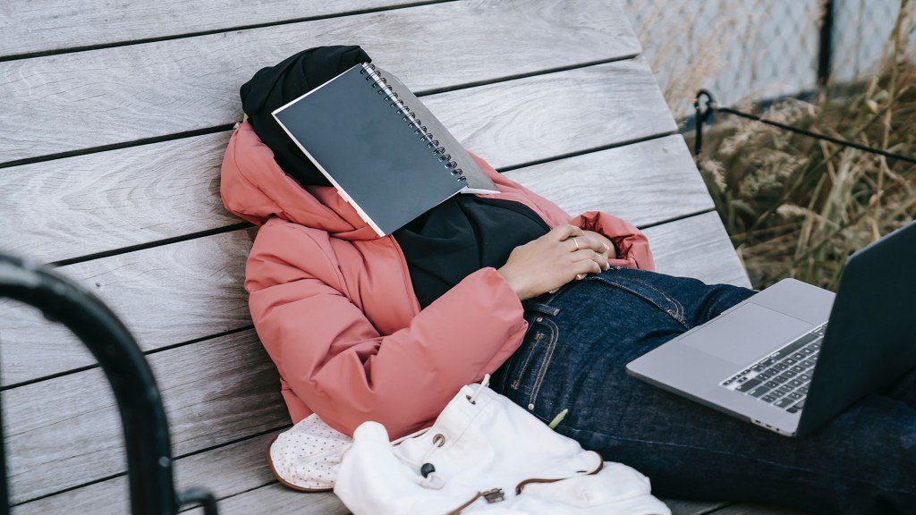 College student laying on bench with a spiral bound book over their face, contemplating whether to add or drop a class.