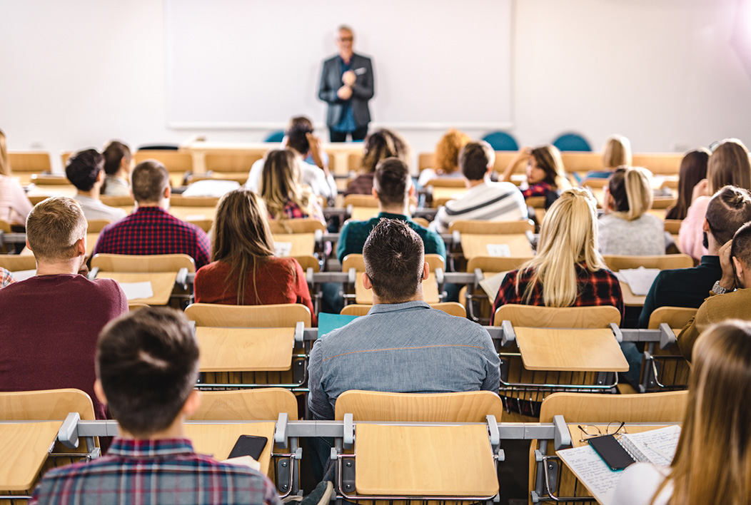 Large classroom full of students, Point-of-View is from rear with teacher at the front.